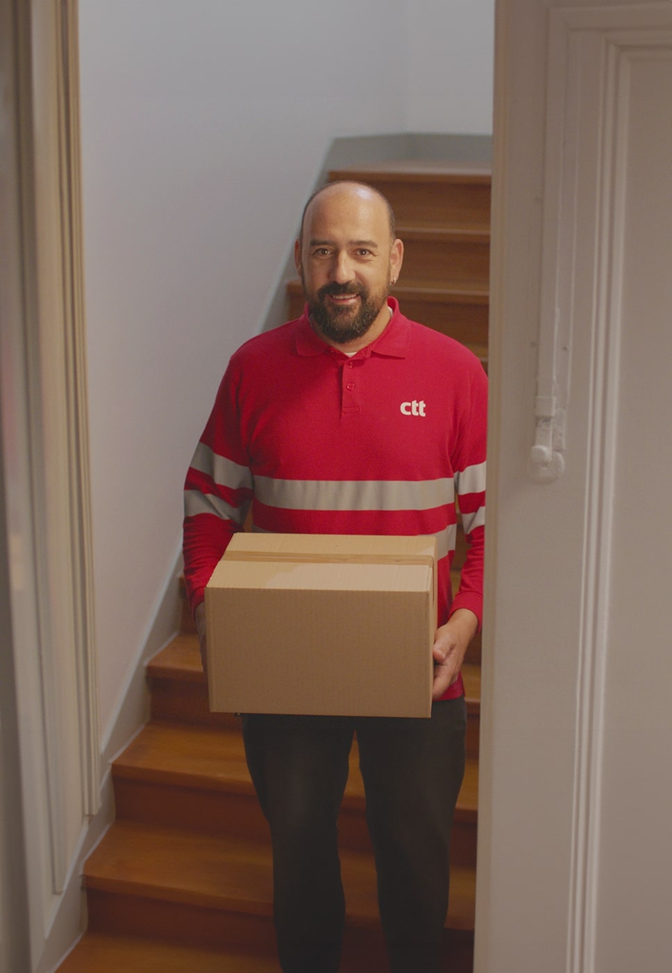 Duas pessoas, uma trabalha ao computador, o outro é carteiro e segura uma caixa de cartão.